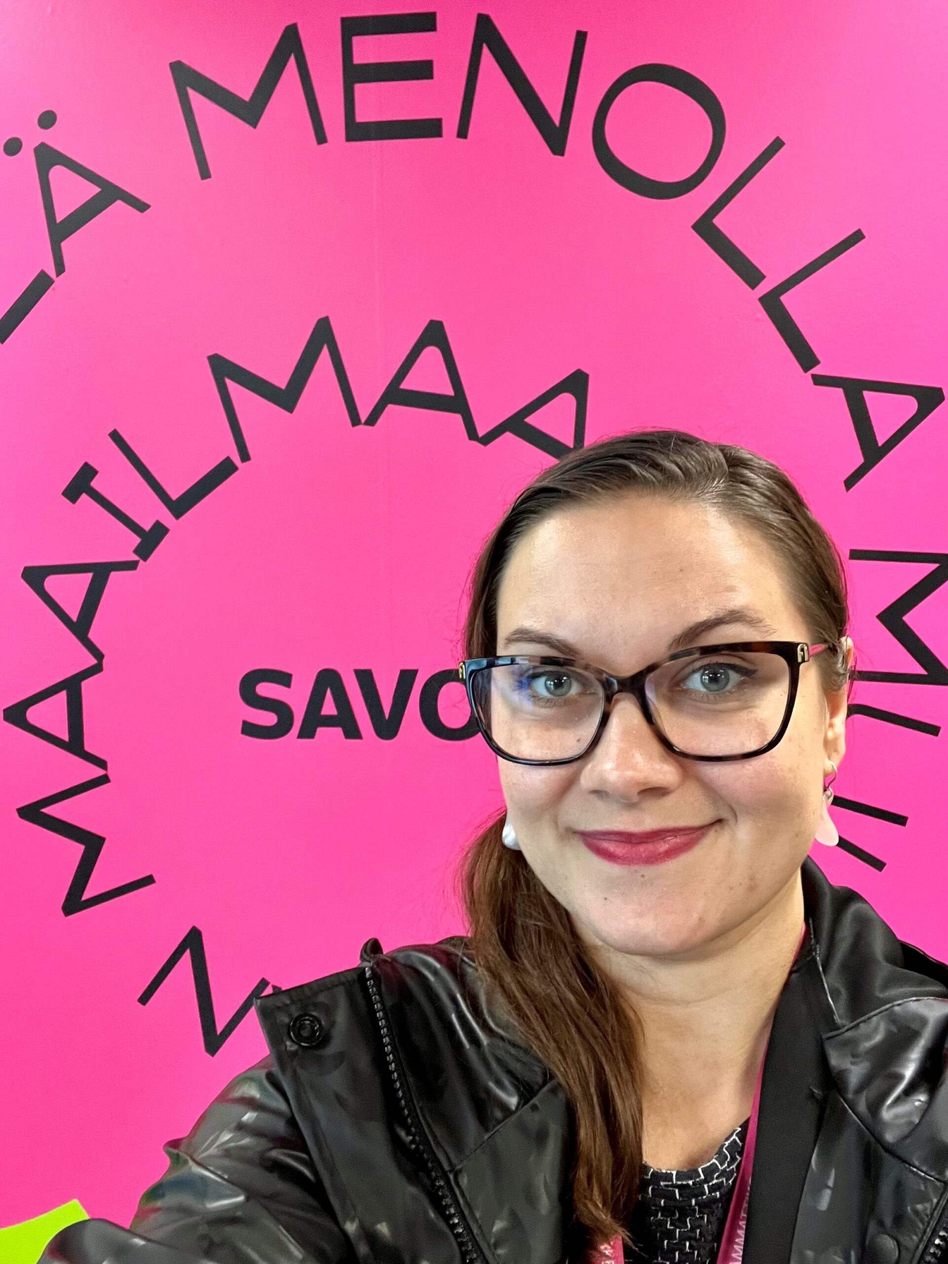 Silmälasipäinen, ruskeahiuksinen nainen hymyilee kirkkaan vaaleanpunaisen seinän edessä, jonka keskellä on suuri, musta ympyränmuotoinen teksti suomeksi ja sana SAVONIA.