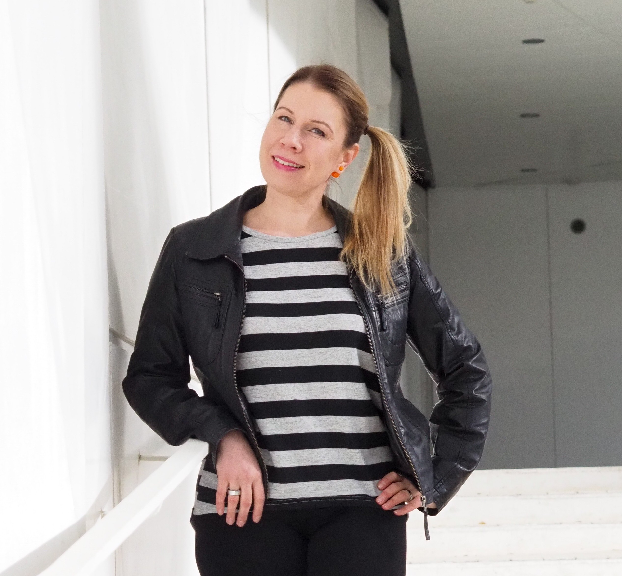 Nainen, jolla on vaaleat hiukset letitettyinä, päällään musta nahkatakki ja mustavalkoraidallinen paita, hymyilee nojaillessaan valkoiseen seinään valoisassa, modernissa sisätilassa.