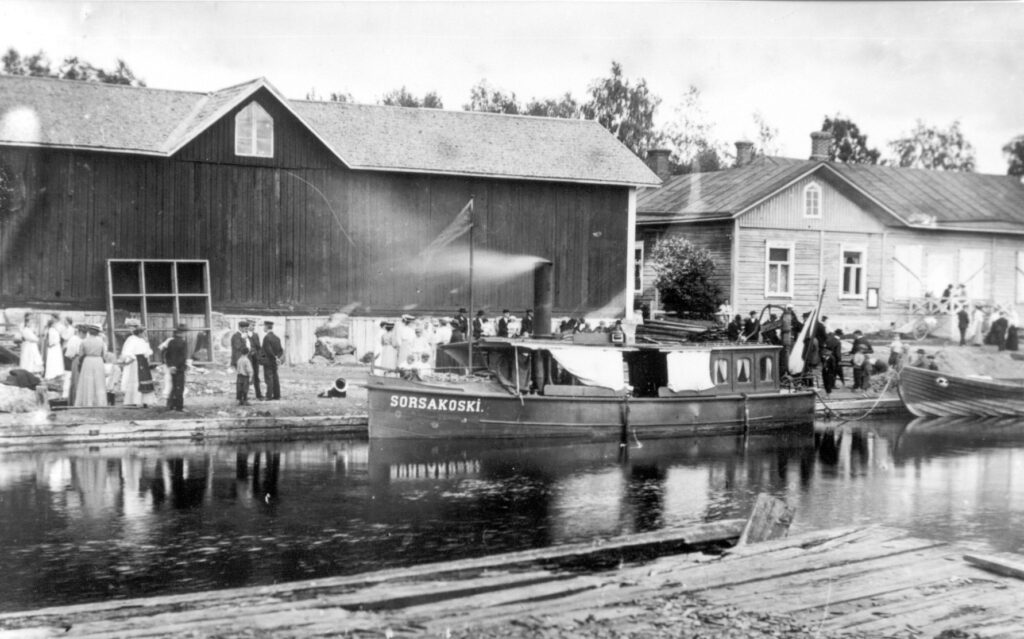 Mustavalkoinen valokuva, jossa Sorsakoski-niminen vene on telakoituna kanavan varrella, ja ihmisiä on kokoontunut rantaan puurakennusten läheisyyteen, todennäköisesti historiallisessa tai 1900-luvun alun ympäristössä.