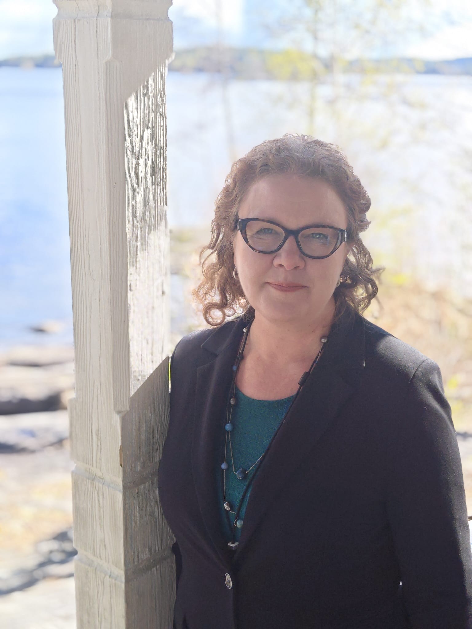 Ruskeat kiharat hiukset ja silmälasit päässä oleva nainen hymyilee valkoisen kuistipylvään vieressä. Taustalla näkyy vesistö ja puita kirkkaalla taivaalla.